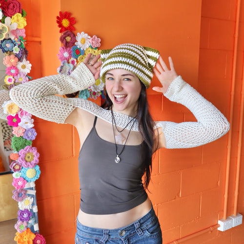 Person wearing a striped crochet hat and white long-sleeve shirt in front of an orange wall with floral decorations.