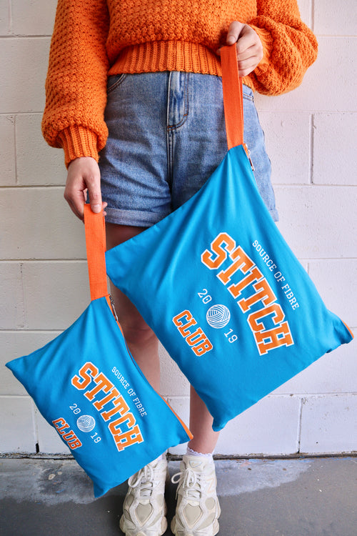 Person holding two blue bags with 'Source Of Fibre Stitch Club' branding against a white wall.